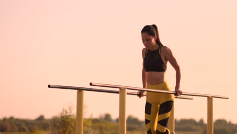 Beautiful-slender-woman-athlete-in-a-black-top-and-yellow-pants-at-sunset-performs-pushups-on-a-parallel-horizontal-bar-in-slow-motion.